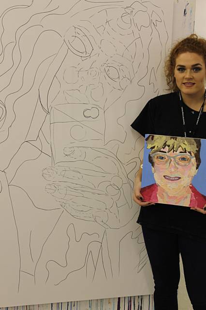 Lois McGee standing in front of a large-scale portrait of a family friend which is she currently painting and holding s smaller, completed piece