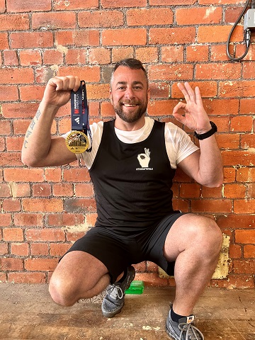  Chris Elliott with his medal. He wears a shirt with the Andy's Man Club logo and is making the 'It's okay to not be okay' sign with his left hand