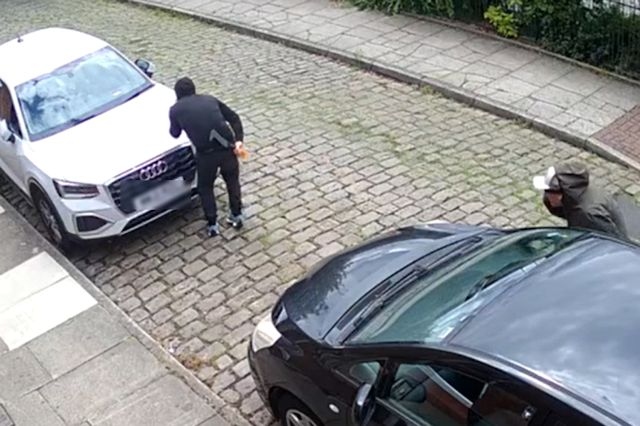 The two men creep around the side of a car, with one clearly holding a billhook machete behind his back