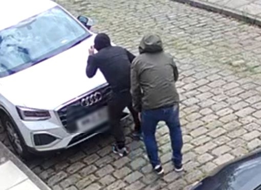 The two men creep around the side of a car, with one clearly holding a billhook machete behind his back